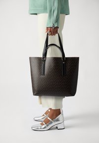 Brun geometriskt mönstrad tote-väska med svarta handtag och guldiga detaljer. Parat med vita byxor och blanka silvriga klackskor.