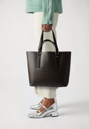 Brown geometric-patterned tote bag with black handles, featuring gold hardware. Paired with white trousers and shiny silver block-heeled shoes.