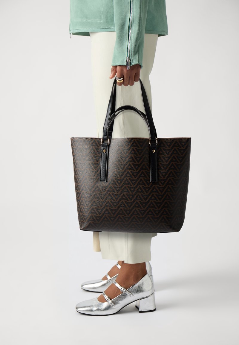 Brun geometriskt mönstrad tote-väska med svarta handtag och guldiga detaljer. Parat med vita byxor och blanka silvriga klackskor.