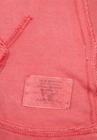 Korallrosa tygmärke med "True Religion"-logotyp, gjort av bomull, och vårdinstruktioner, sydd i plagget med kontrasterande tråd.