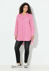 Vrouw met krullend donker haar, gekleed in een lange roze-wit gestreepte blouse, zwarte skinny jeans en witte sneakers, staand tegen een witte achtergrond.