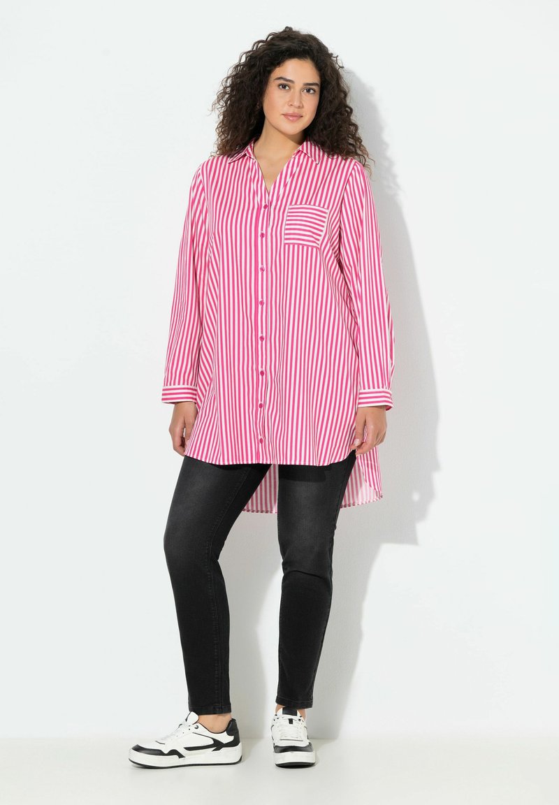 Vrouw met krullend donker haar, gekleed in een lange roze-wit gestreepte blouse, zwarte skinny jeans en witte sneakers, staand tegen een witte achtergrond.