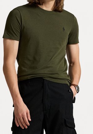 Jeune personne portant une chemise à manches courtes de couleur vert olive avec un petit logo brodé et un pantalon cargo noir, main dans la poche.