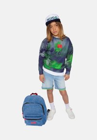 Zaino in denim blu con marchio Hugo accanto a un bambino che indossa una felpa a motivi blu navy e verde, pantaloni in denim e sneaker bianche.