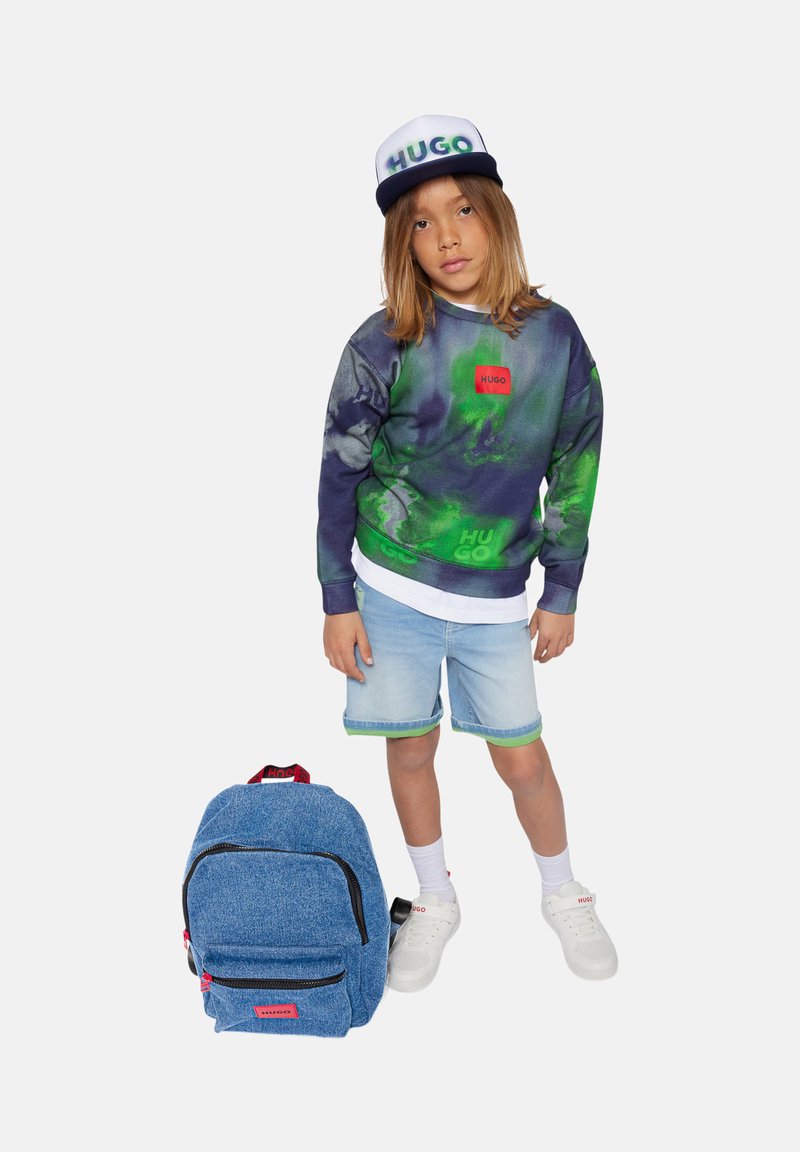Zaino in denim blu con marchio Hugo accanto a un bambino che indossa una felpa a motivi blu navy e verde, pantaloni in denim e sneaker bianche.