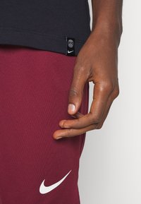 Czarna koszulka z małą naszywką z logo oraz bordowe spodnie z białym logo Nike w kształcie łyżwy. Gładka tkanina o dopasowanym kroju. Widoczna ręka.