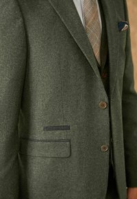 Veste de costume en mélange de laine verte avec une finition texturée, revers cranté et fermeture à deux boutons. Comprend une poche poitrine avec un mouchoir à motif.