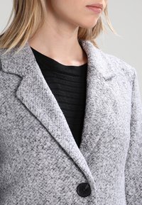 Šedý strukturovaný vlněný kabát smíchaný s jinými materiály, se zářezovým límcem a jedním černým knoflíkem, přelité přes černý ribbed top.