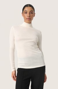Long-sleeve turtleneck shirt in cream, made of lightweight fabric. Features a fitted design and smooth texture, paired with black trousers.