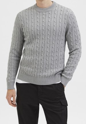 Man wearing a light grey cable knit sweater over a white shirt, paired with black cargo pants, standing with one hand in pocket.