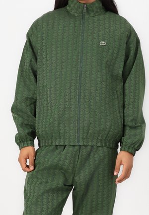 Mann trägt eine grün gemusterte Zip-up-Jacke und passende Hosen mit einem gestickten Krokodil-Logo auf der Brust.