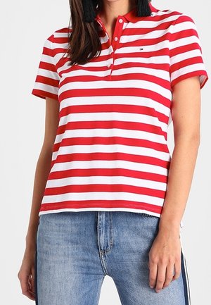 Kobieta ubrana w koszulkę polo z krótkim rękawem w czerwono-białe poziome paski, w połączeniu ze średniozabarwionymi niebieskimi dżinsami.