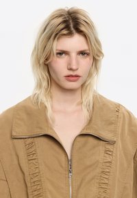 Una giovane donna con capelli biondi spettinati e trucco minimal indossa una giacca marrone con zip e dettagli arricciati, guardando direttamente verso la fotocamera.