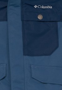 Veste bleue avec une poche à rabat bleu foncé maintenue par un bouton pression et un logo Columbia blanc au-dessus de la poche.
