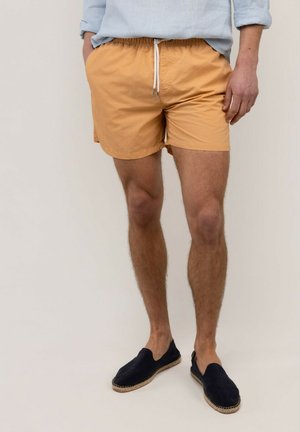 Hombre con camisa azul clara remangada, pantalones cortos color canela con cordón blanco y zapatos negros sin cordones, de pie sobre fondo liso.