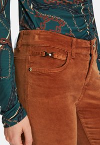 Braune Cordhosen weisen einen leichten Glanz, einen schmalen Schnitt, Fronttaschen mit einem Metallelement und eine strukturierte Oberfläche in einem warmen Farbton auf.