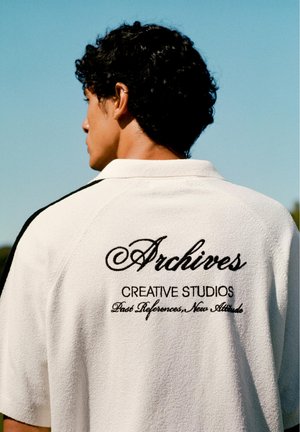 Jeune homme aux cheveux courts et bouclés portant une chemise blanche avec le texte « Archives Creative Studios, Past References, New Attitude » dans le dos.