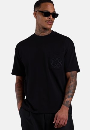 Mann trägt schwarzes T-Shirt mit Tasche und dezentem geometrischem Muster, schwarze Hose, Sonnenbrille und sichtbare Tattoos am Hals und an den Armen, Hände in den Taschen.