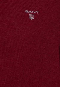 Tissu en tricot bordeaux avec le logo GANT et un emblème en forme de bouclier brodé près du centre supérieur.