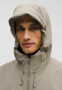 Chaqueta beige con capucha de textura suave, cuello alto y cierre de cremallera completo. Presenta cordones ajustables en la capucha para un ajuste seguro.