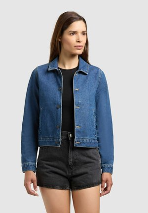 Frau mit langen braunen Haaren, die eine blaue Jeansjacke mit Knöpfen über einem schwarzen Top und hoch taillierte schwarze Jeansshorts trägt.