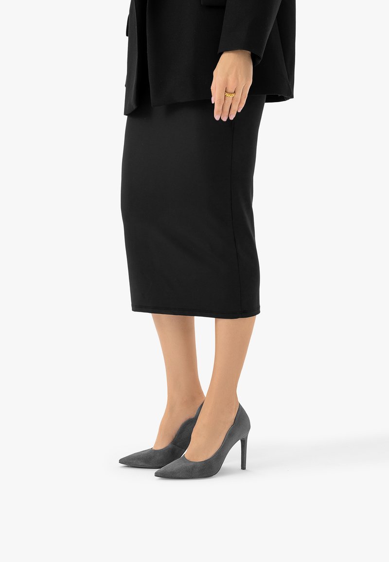 Femme portant une jupe crayon noire, un haut noir et des chaussures à talons gris, montrant une main avec une bague en or sur fond blanc.
