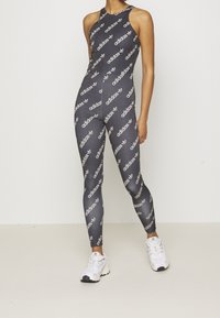 Czarny sportowy kombinezon z białym nadrukiem logo adidas, z wysokim kołnierzem i dopasowanym krojem. Połączony z białymi sneakersami.