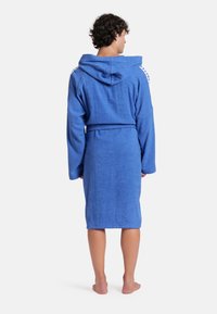 Blauwe badjas van badstof met capuchon, vastgebonden in de taille. Heeft lange mouwen en contrasterende zijaccenten. Zachte textuur en knielange snit.