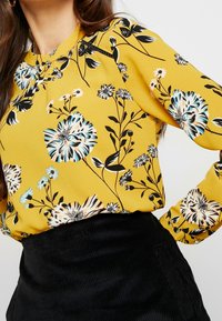 Donna che indossa una camicetta floreale gialla con fiori neri, bianchi e blu, infilata in una gonna nera di velluto a costine a vita alta.