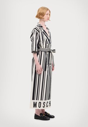 Robe rayée noir et blanc à manches courtes, avec une ceinture à la taille et des poches. L'ourlet comporte le texte "MOSCH." Portée avec des mocassins noirs.