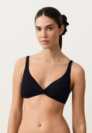 Nainen, jolla on tummat hiukset sidottuna takaa hiusdonitsilla, pukeutuneena mustaan V-kaulaiseen braletteen, jossa on etuosassa kiertynyt kangasdetaili.