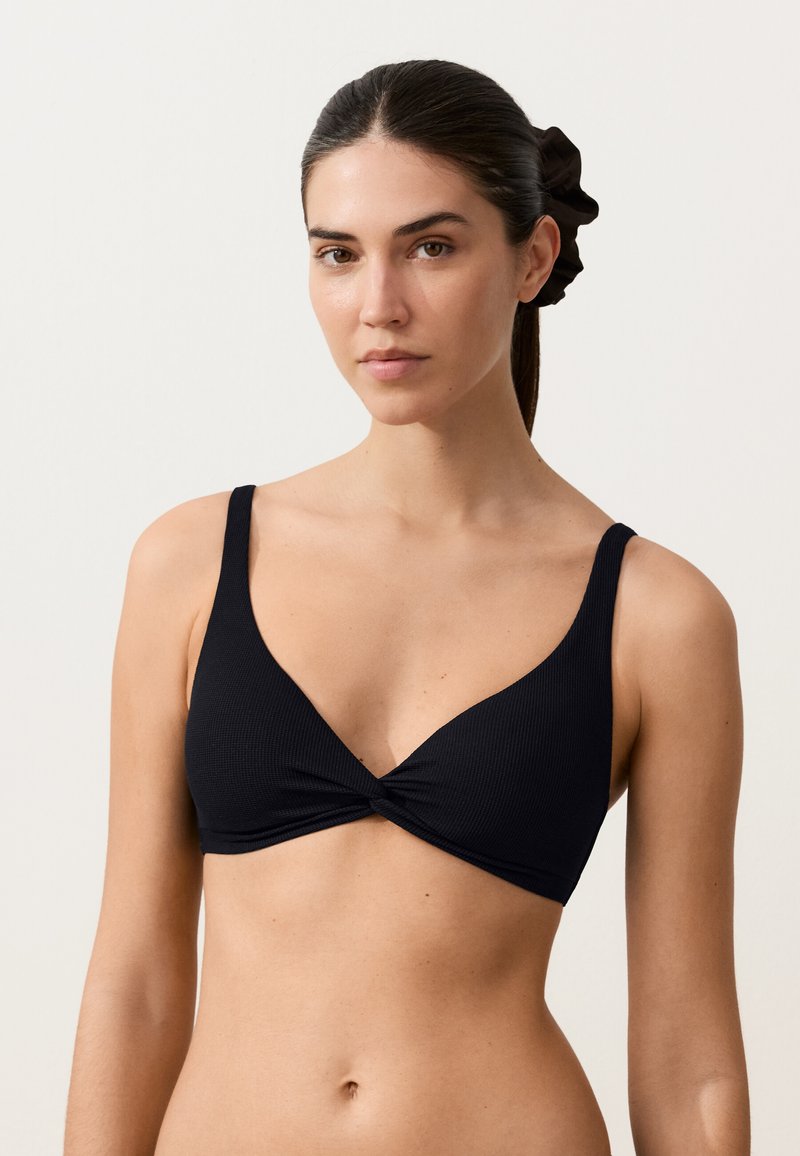Femme aux cheveux foncés attachés en arrière avec un chouchou, portant un bralette noir en V avec un détail de tissu torsadé à l'avant.
