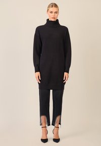 Schwarzer Strickpullover mit Rollkragen und langen Ärmeln, kombiniert mit schwarzen Hosen mit Fransenabschlüssen, die mit Perlen verziert sind, und hohen Schuhen.