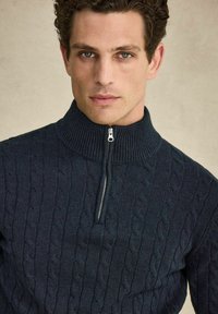 Mörkblå stickad tröja med hög krage, dragkedja och mönster i strukturstickning. Mjukt material med ribbade detaljer.