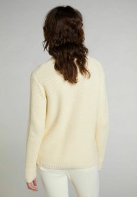 Kermanvärinen, teksturoitu neulepusero, jossa pitkät hihat ja pyöreä pääntie. Löysä istuvuus ja resorit helmassa, nähtynä takaa.