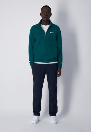 Giacca zip-up color turchese con logo bianco, indossata sopra una camicia bianca, abbinata a pantaloni felpa blu navy e scarpe da ginnastica bianche. Tessuto morbido, vestibilità casual.