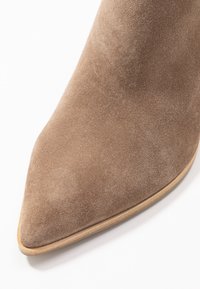 Kärkkäät nilkkurit, jotka on tehty taupe-värisestä mokasta. Sujuva pinta, jonka muotoilu on tyylikäs ja beige-pohja on hienostunut. Minimalistinen design, jossa ei ole näkyvää metalliosaa.