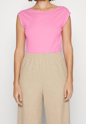 Frau trägt ein leuchtend pinkes ärmelloses Oberteil und weite beige Hose mit hohem Bund und elastischem Taillenbund, steht vor weißem Hintergrund.