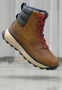 Bottine de randonnée en cuir marron avec un col en polaire grise, des lacets rouges et bleus, et une semelle en caoutchouc noir. Présente un logo sur le côté et une languette rembourrée.