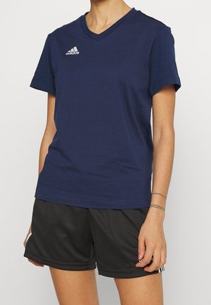 Person iført en navyblå Adidas V-hals T-shirt og sorte sports-shorts, stående mod en ensfarvet lys baggrund.