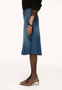 Person som bär en midilång denimkjol, svarta genomskinliga strumpbyxor, bruna slingbacksandaler, svart topp och tjocka armband på vänster handled.