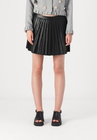 Jupe plissée en simili cuir noir avec une taille lisse, associée à des sandales noires ouvertes, mettant en avant un design épuré et minimaliste.