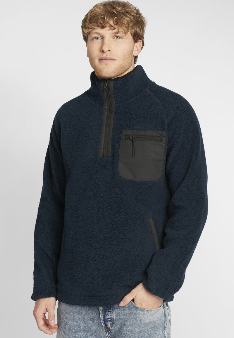 Navy fleece pullover with a half-zip collar, featuring a black zippered chest pocket and side pockets, textured fabric, and relaxed fit.