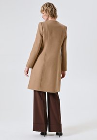 Lungo cappotto di lana beige con taglio dritto, cuciture definite e maniche lunghe, abbinato a pantaloni larghi marroni. Sfondo semplice.