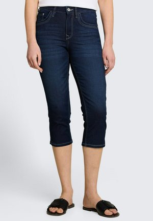 Frau trägt dunkelblaue kurze Jeans und schwarze Slip-On-Sandalen vor einem einfarbigen hellen Hintergrund.