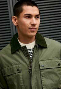 Olivgrüne Jacke mit einem Cordkragen, Reißverschluss und zwei Brusttaschen. Darunter ist ein cremfarbenes Sweatshirt mit einem schwarzen Muster sichtbar.