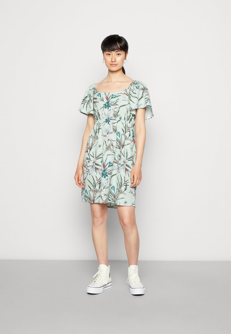 Vestido com padrão floral em verde claro, com mangas curtas e soltas, cintura ajustada e comprimento até o joelho, combinado com ténis brancos de cano alto.