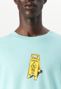 Light blue cotton t-shirt featuring a yellow graphic of a caution sign, labeled "DANCE FLOOR," on the front.