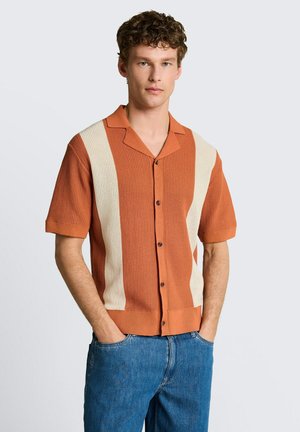 Młody mężczyzna w pomarańczowo-rdzawej i kremowej dzianinowej koszuli z krótkim rękawem i guzikami, w niebieskich jeansach, stoi z rękami w kieszeniach na jednolitym tle.