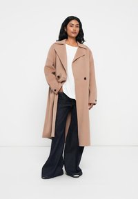 Cappotto beige in misto lana con revers a punte, bottoni doppi e una vestibilità rilassata, abbinato a pantaloni flare in denim scuro.